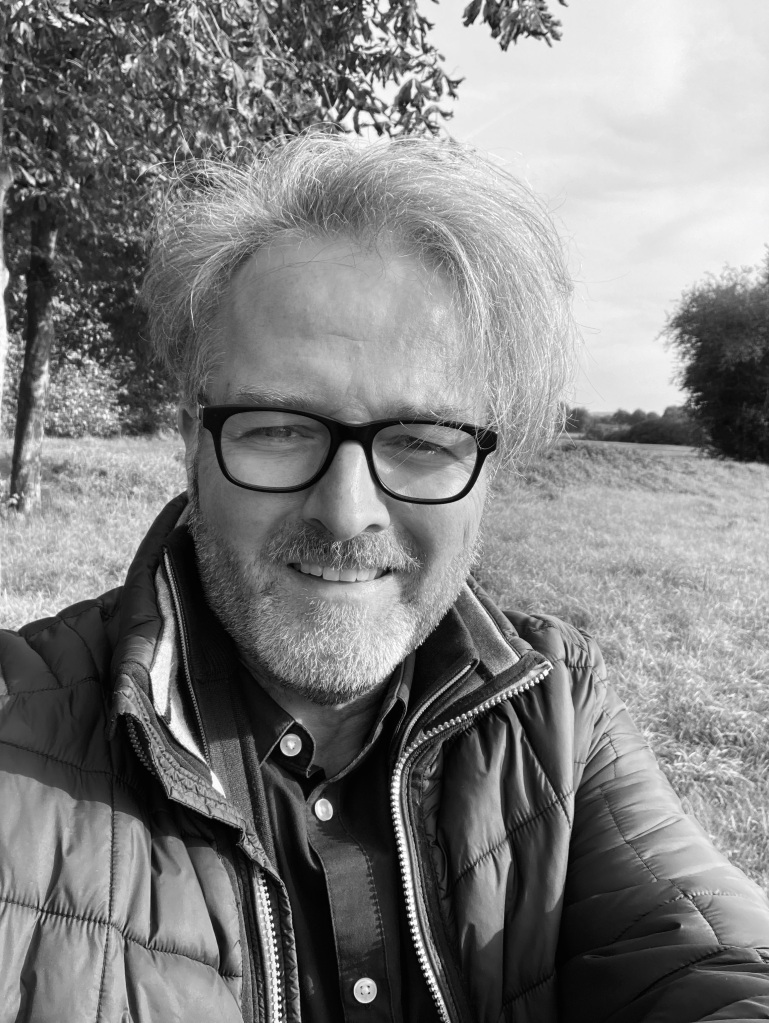 Schwarz-weißes Selfie von Stefan Remeke in Hemd und Übergangsjacke vor Feldlandschaft mit Bäumen.